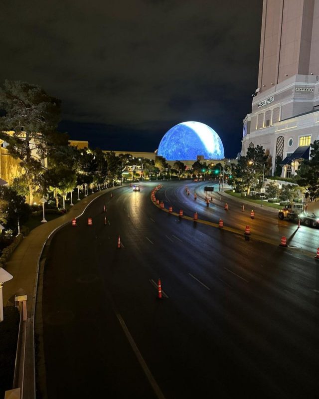 Night time cityscape of an empty road lined with traffic cones. The road is damp from recent rain.